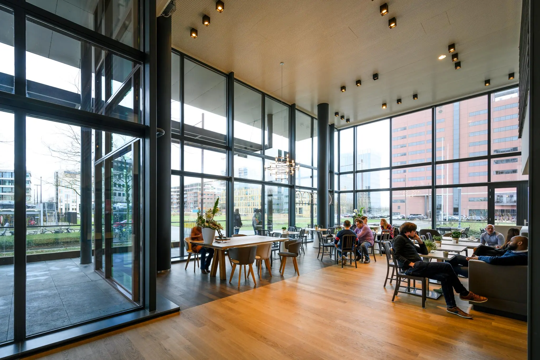 Moderne en ruime ontmoetingsruimte met grote ramen aan de Thomas R. Malthusstraat, waar mensen ontspannen en met elkaar praten.